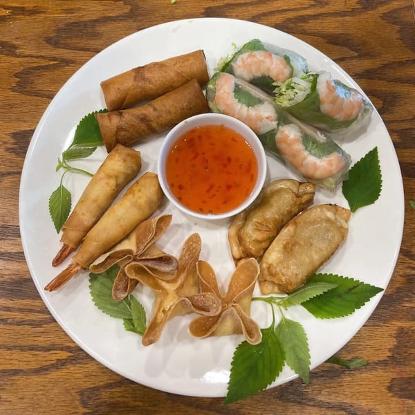 A white plate of Vietnamese appetizers including spring rolls, shrimp rolls, and crab rangoons with dipping sauce.